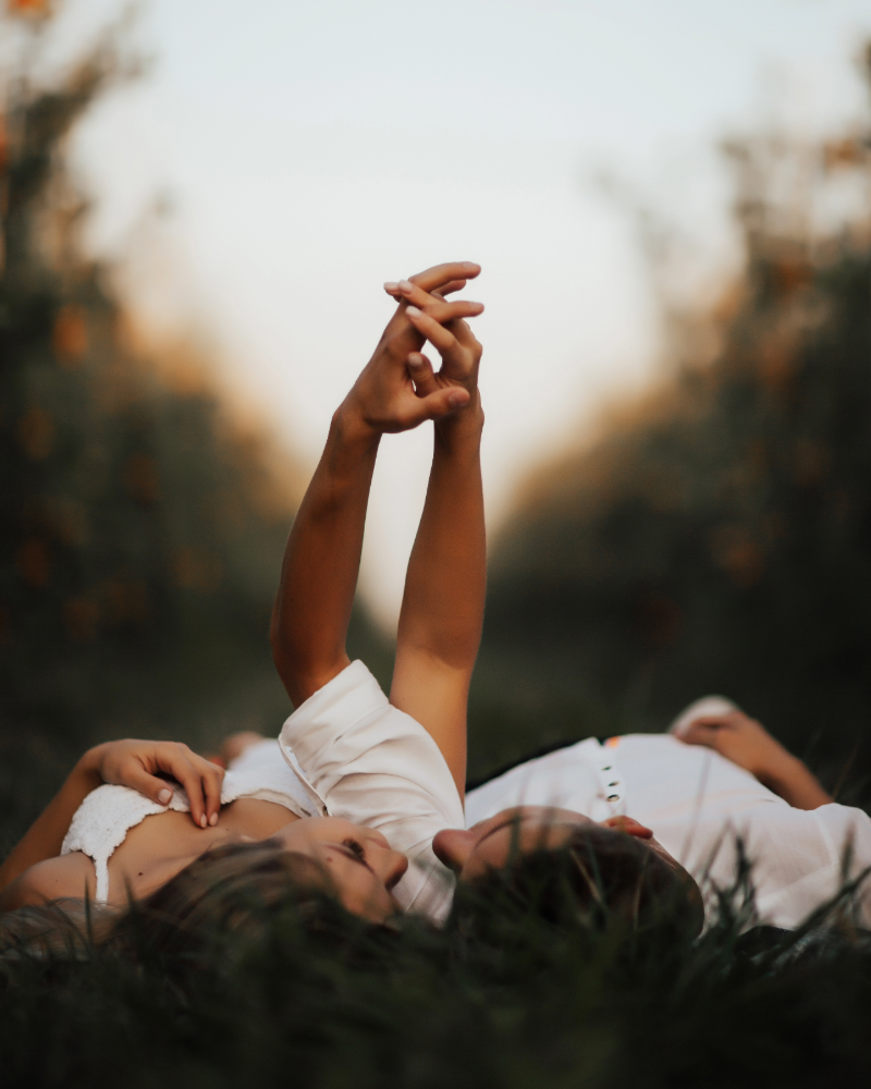 Couple lying down outside, in the dusk. Both arms up in the air outstretched with fingers intertwined. Both dressed in white summer wear