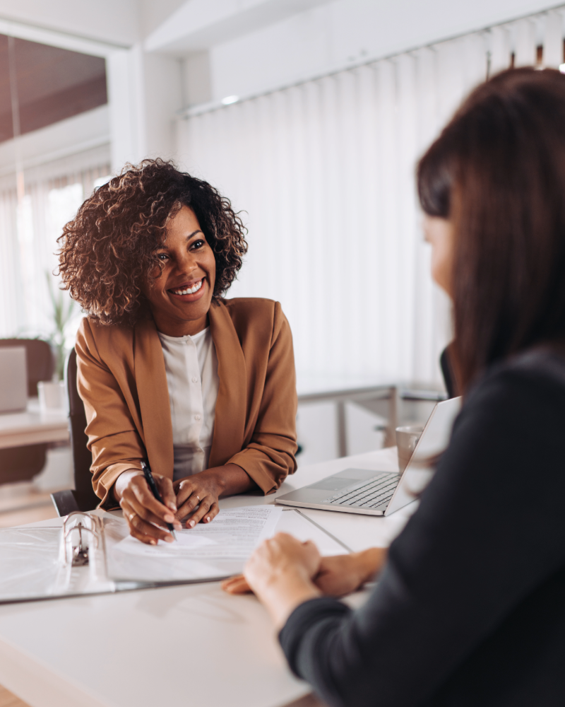 two women sat at a desk. one facing the camera smiling, has an afro. One facing away from the camera, dark long frown hair. Laptop and paper on the desk. white vertical blinds in the background. fertility consultant